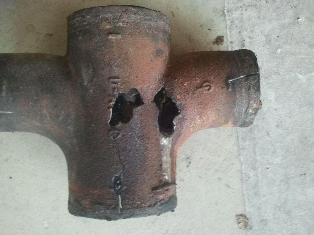 A rusted cast-iron cross fitting with two holes corroded through its wall, lying on concrete