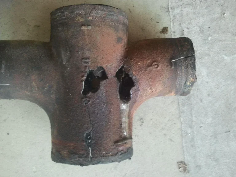 A rusted cast-iron cross fitting with two holes corroded through its wall, lying on concrete