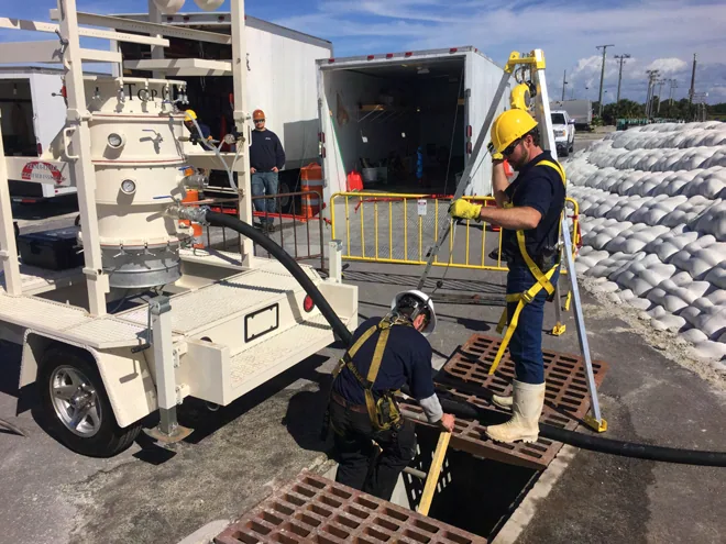 Blue Works crew in hard hats and fall-protection harnesses at an open drain grate at the FPL industrial drain